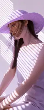 Nainen, joka käyttää leveälieristä violetin väristä olkihattua ja vaaleanvärisiä vaatteita.